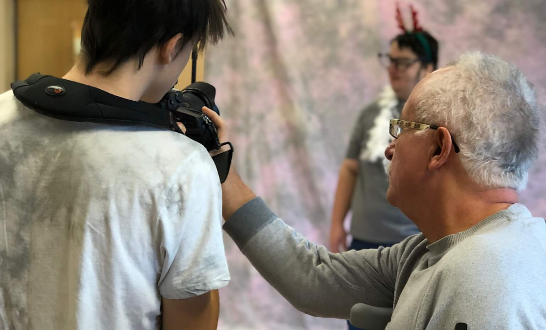 Teacher holding a camera for a student and teaching them to photograph another student on a backdrop.